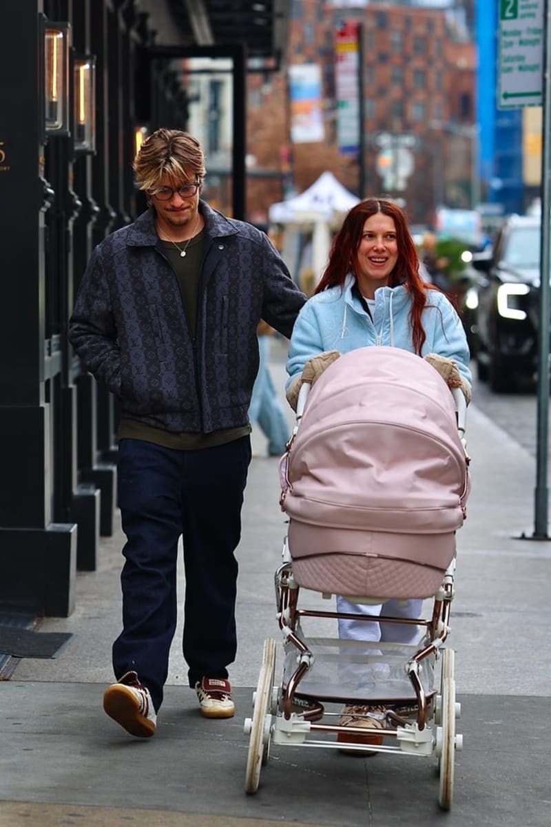 A man in a monogram jacket and a woman in a light blue hoodie walk along a city pavement pushing a large pink pram.