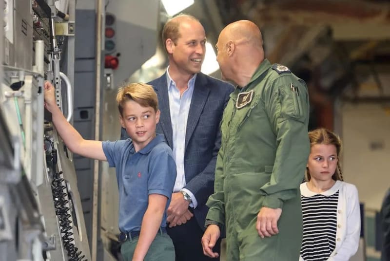 A man in a blue blazer stands with a young boy and girl inside a military aircraft, alongside a bald officer in a green flight suit.