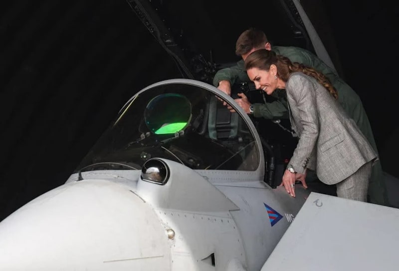 A woman in a grey blazer with a braided ponytail leans over the cockpit of a military jet alongside a pilot in a green flight suit.
