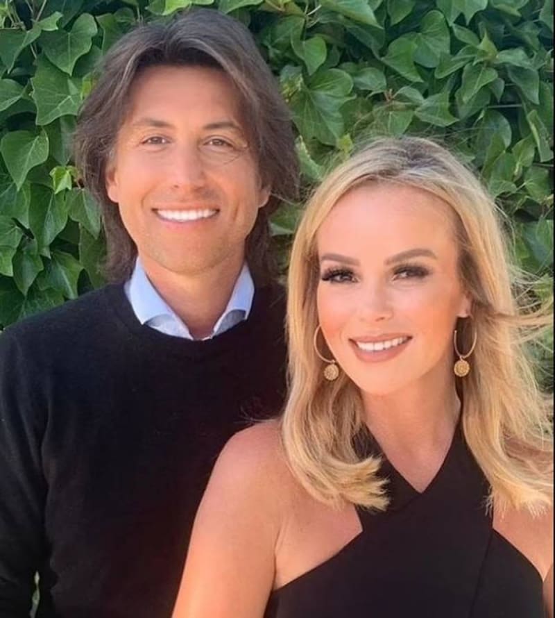 Two people smiling at the camera outdoors with green foliage in the background.