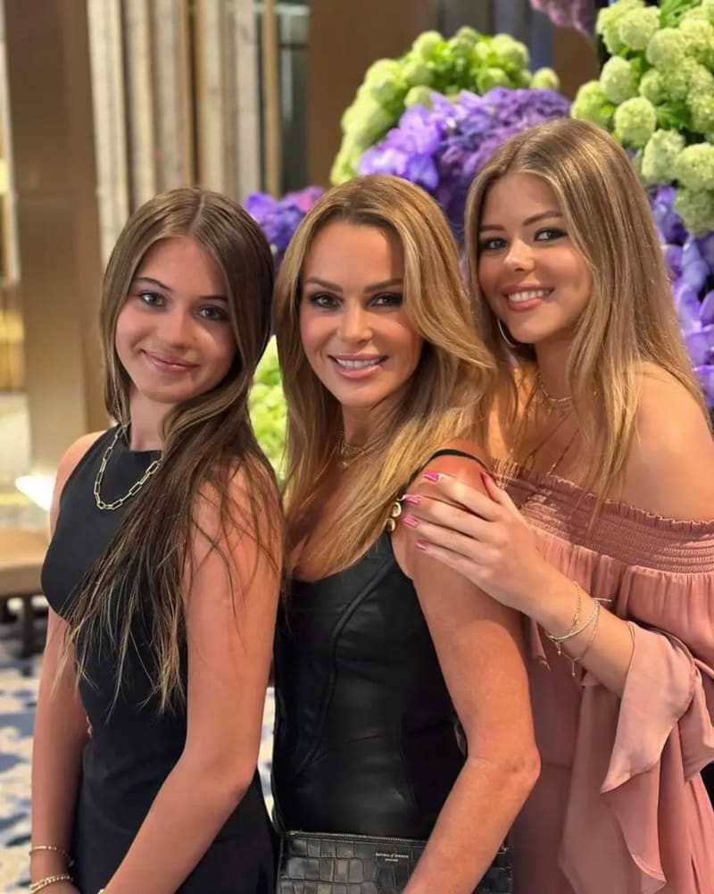 Three women with long brown hair pose together in front of a floral arrangement — the woman in the centre wearing a black leather dress, flanked by two younger women in black and pink respectively.