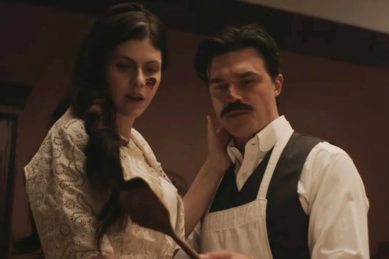 A woman in a lace-trimmed blouse and a man with a mustache wearing a vest and apron stand together indoors.