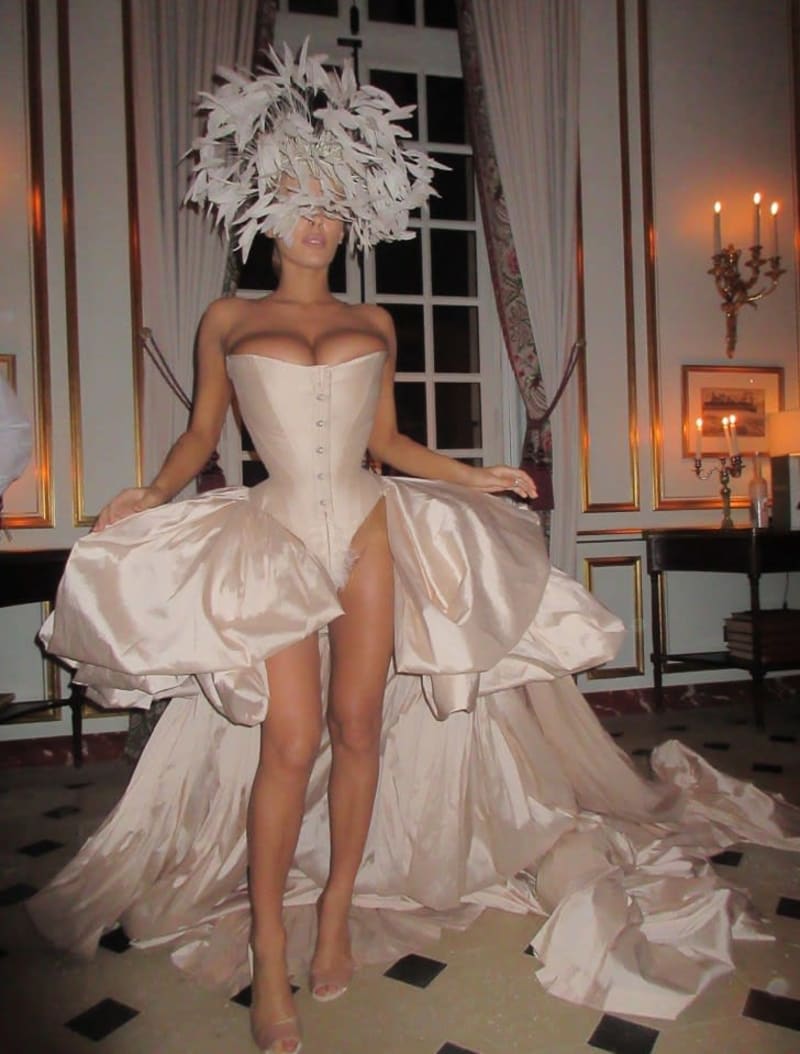 A woman in a white strapless corset dress and large feathered headpiece poses in an elegant room with chandeliers.