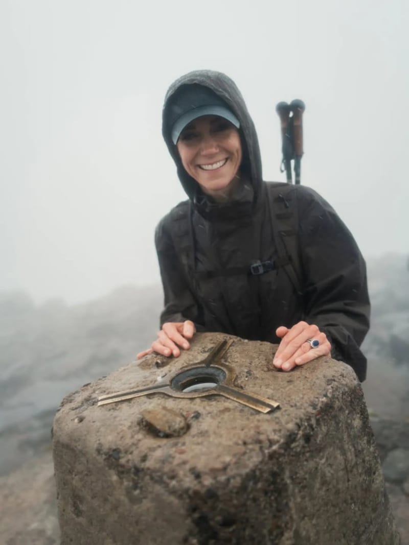 A woman in a black waterproof jacket and cap leans over a stone trig point on a misty mountain summit, wearing a sapphire and diamond ring, with walking poles visible behind her.