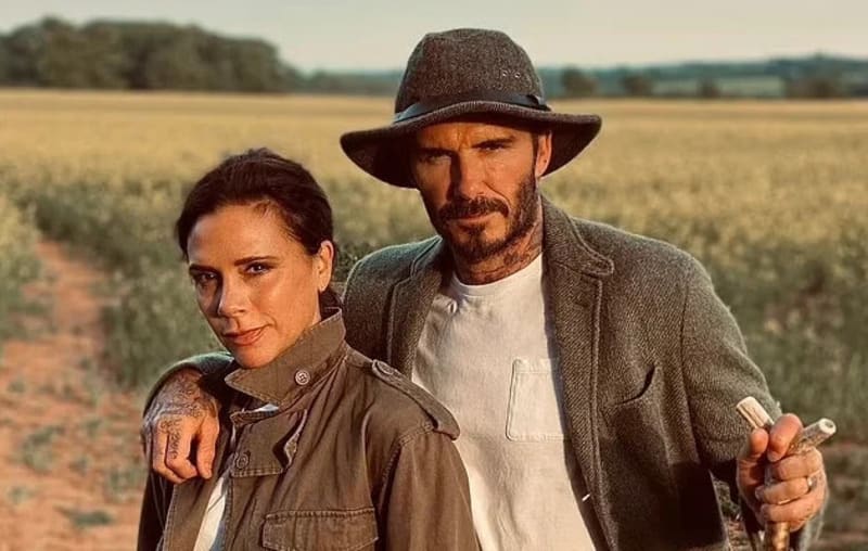 A man and woman in rustic outfits against backdrop of a farm field. Man in hat, leather jacket white t-shirt, woman in brown leather jacket over white t-shirt