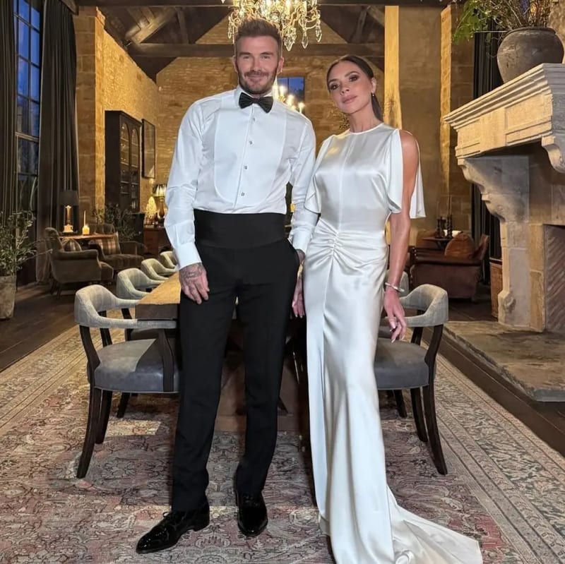A man in a white dress shirt and black bow tie stands beside a woman in a white satin gown inside a stone-walled manor house with a chandelier, fireplace and candlelit dining room behind them.