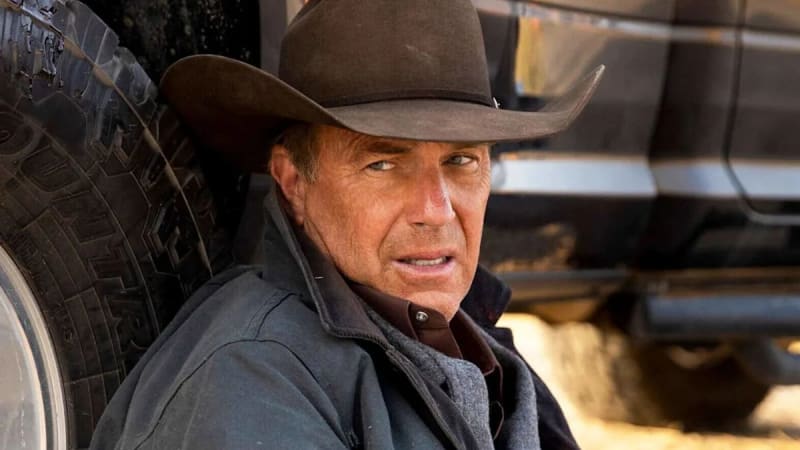 A man wearing a brown cowboy hat and dark jacket leans against a large truck tire in an industrial setting.