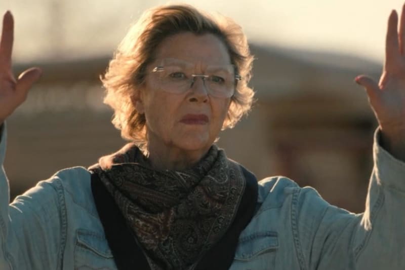 A woman wearing glasses and a denim jacket with a black bandana looks upward with raised hands in an outdoor setting.