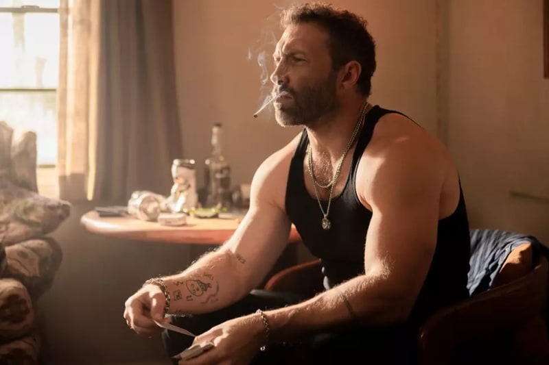 A bearded man with tattoos smoking a cigarette in a sunlit room, wearing a black tank top and chain necklace.