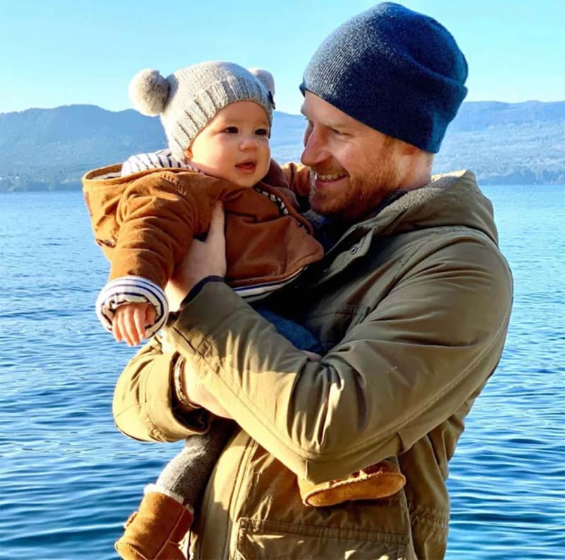 A bearded red-haired man in a green parka and blue beanie holds a baby in a tan padded jacket and grey pom-pom hat, smiling at the child against a blue lake and mountain backdrop.