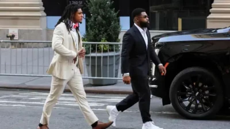 Two men walk across a city street — one in a cream suit with a pink bow tie and brown brogues, the other in a dark suit with sunglasses and white trainers, a large black SUV beside them.