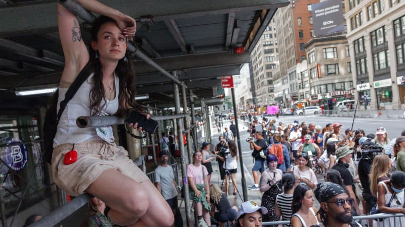A woman with a tattoo sits on an elevated platform above a crowded urban street event, holding binoculars and looking into the distance.