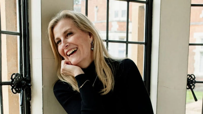 A blonde woman in a black turtleneck laughs with her chin resting on her hand, leaning against a window frame with a brick building visible behind her.