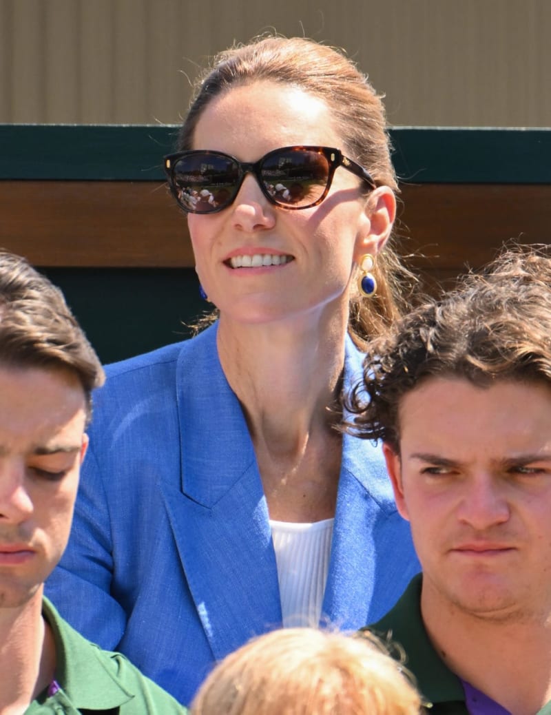 A woman wearing a blue blazer and dark sunglasses smiles while seated among other spectators at an outdoor event.