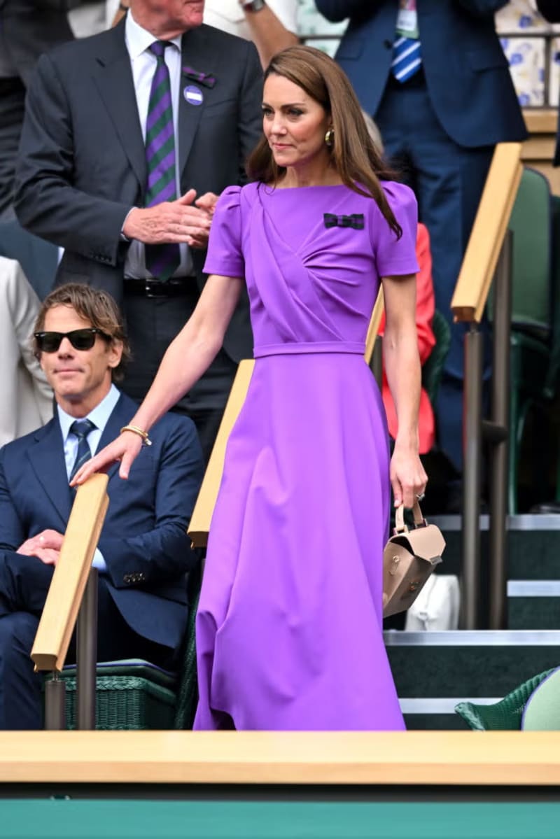 A woman in a purple short-sleeved A-line dress with a black bow detail makes her way through the royal box seating at Wimbledon, holding a small tan bag.