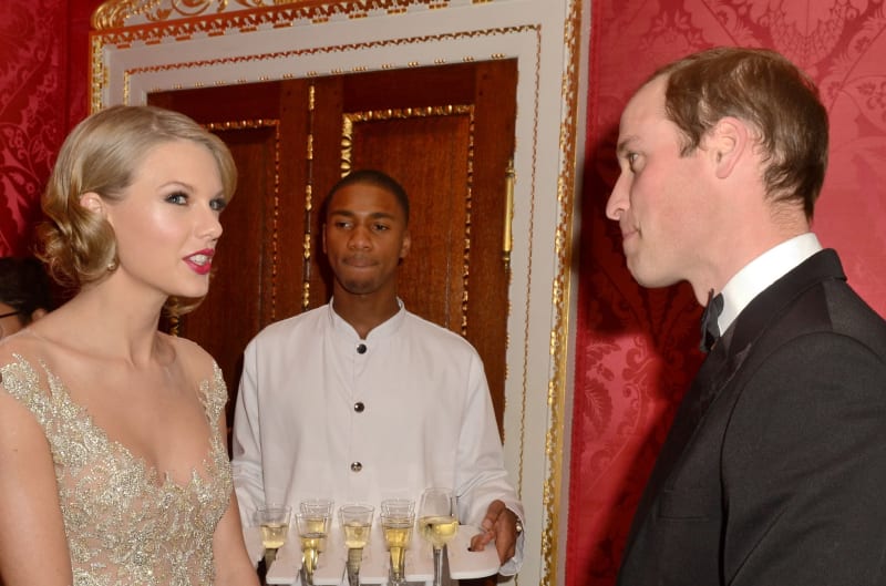 Three people at a formal event in an ornate red room with gold-trimmed doors, with champagne glasses on a table.