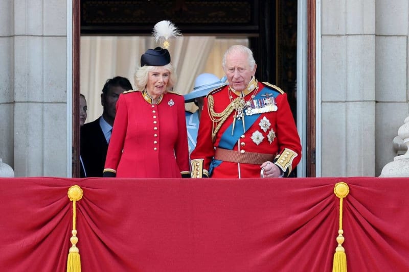 Two people in formal red ceremonial dress stand on a palace balcony behind a red and gold draped railing.