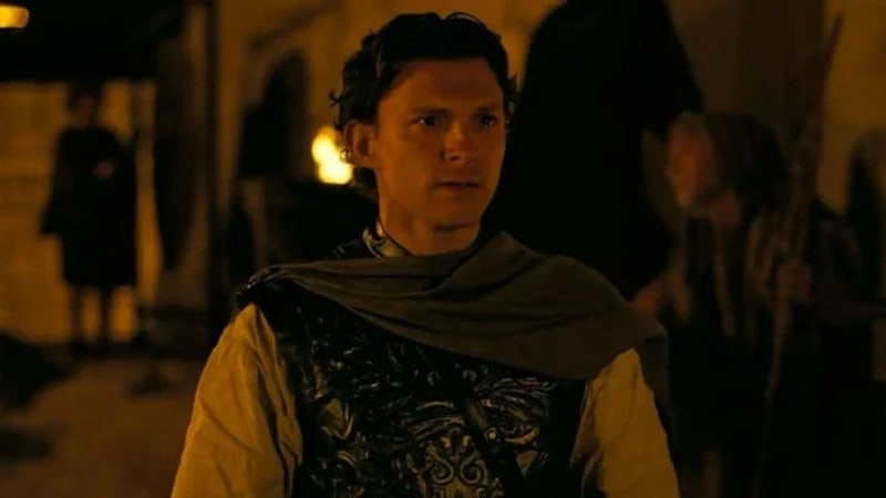 A man in medieval clothing with a gray cloak and ornate black vest stands in a dimly lit street scene.