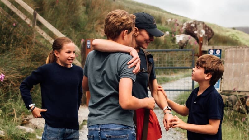 A man in a gray shirt and baseball cap embraces a woman while two children stand nearby in a rural garden setting.