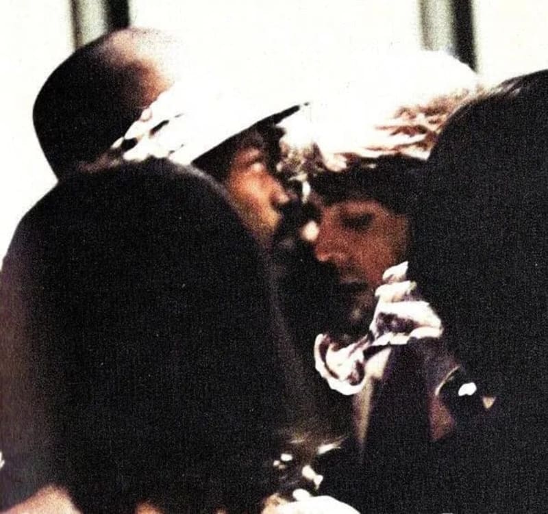 Close-up of two people embracing closely at a low angle, faces obscured by curly and dark hair, in a grainy, low-resolution photograph.