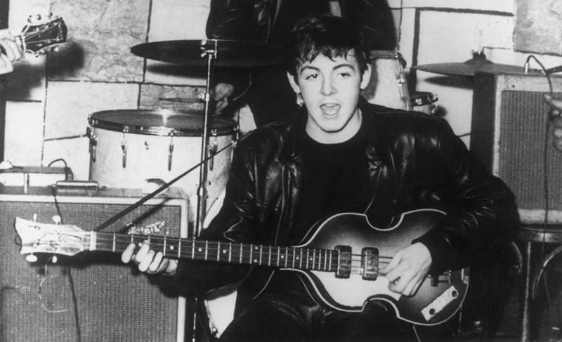 A young man with tousled dark hair in a leather jacket plays a violin-shaped bass guitar and sings, with a drum kit and amplifiers behind him in a stone-walled room, in a black-and-white photograph.