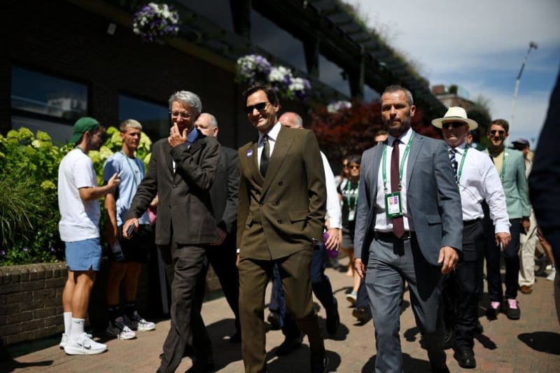 A group of formally dressed men walk together at an outdoor event, with spectators and hanging flowers visible in the background.