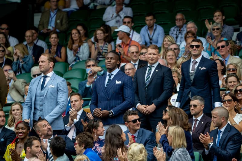 Four men in tailored suits stand together in a crowd, applauding, at a tennis stadium.