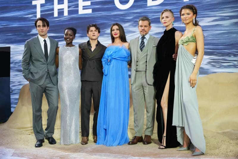Seven people standing in a row on a sandy-themed stage backdrop with ocean imagery and partial text reading 'The Od—'; from left to right: a man in a grey suit, a woman in a silver sequined gown, a young man in a dark vest and trousers, a woman in a voluminous blue off-shoulder gown, a man in a tan three-piece suit, a woman in a black sleeveless dress with a high slit, and a woman in a sage green draped gown with a cutout midriff.