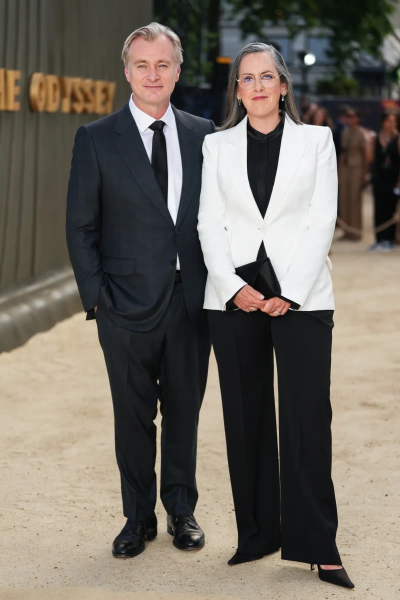 A man in a dark navy suit with a black tie stands beside a woman in a white blazer over a black blouse and wide-leg trousers, holding a black clutch, on a sandy outdoor premiere carpet with a partial golden 'The Odyssey' sign visible behind them.