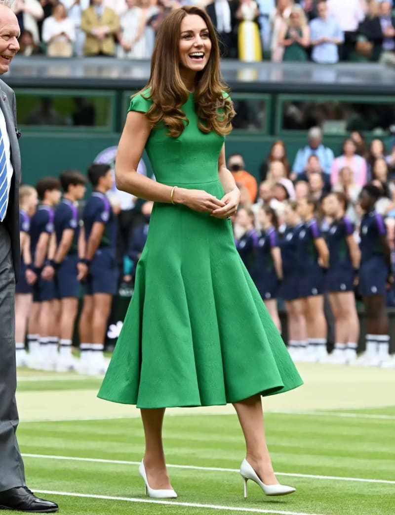 A woman in a bright green short-sleeved flared midi dress walks on a grass court, laughing, with ball boys and girls in the background.