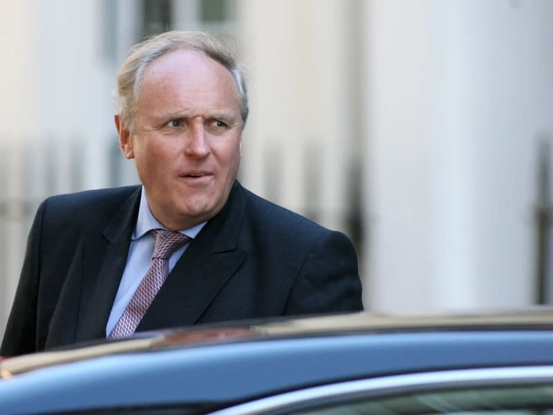 A middle-aged white man with grey-blond hair wearing a dark suit jacket, light blue shirt, and patterned tie, standing outdoors near a dark car with a white columned building in the background.
