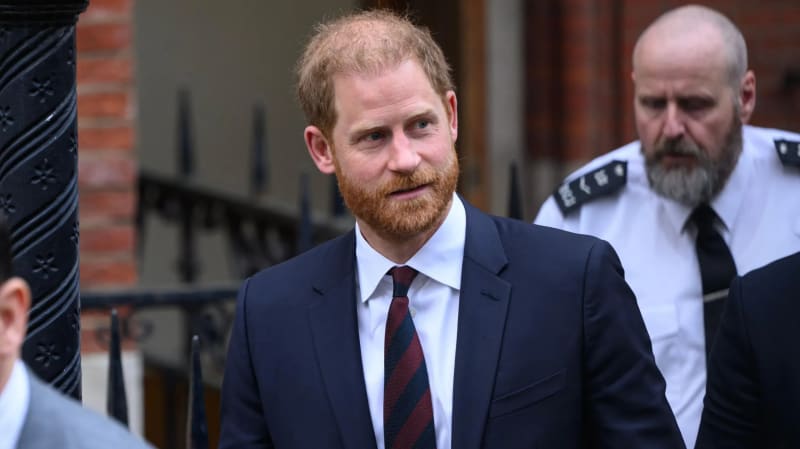 A red-haired, bearded man in a navy suit and striped burgundy tie stands outdoors near a stone building entrance, with a bald, bearded man in a white uniform shirt and black tie visible behind him.
