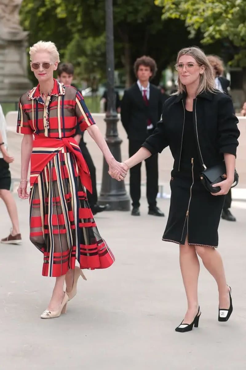 Two women walking hand-in-hand on a paved outdoor path, the woman on the left wearing a sheer red plaid midi dress with nude heels and short platinum hair, and the woman on the right wearing a black zip-detail jacket and skirt with black slingback heels and carrying a small black clutch, with a young man in a suit visible in the background.