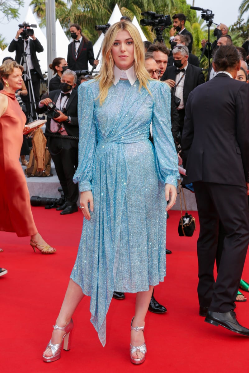 A young blonde woman stands on a red carpet wearing a long-sleeve light blue sequined midi dress with a white collar and silver platform heels, surrounded by photographers and formally dressed attendees in the background.