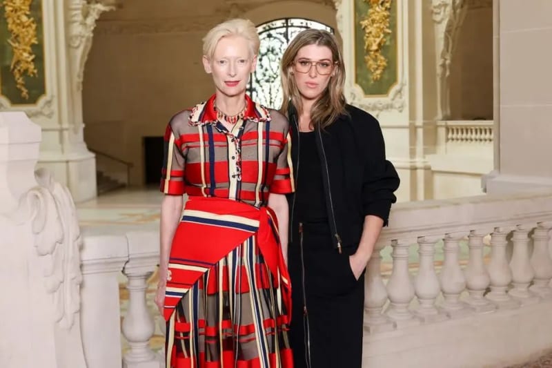 Two women standing together in an ornate white and gold interior with marble balustrades, one wearing a bold red plaid and striped midi dress with sheer detailing and short blonde hair, the other in an all-black outfit with glasses and shoulder-length brown hair.