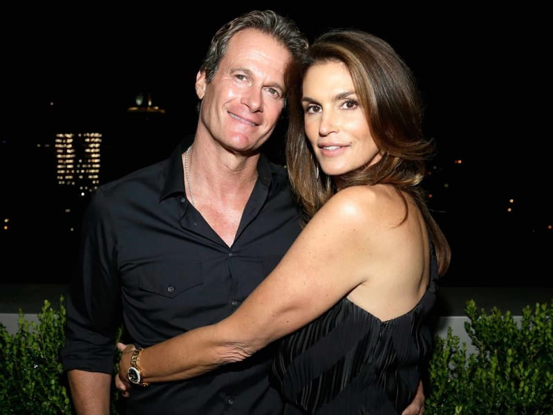 A man in a black button-down shirt with a gold watch and chain necklace stands with his arm around a woman in a black sleeveless dress, both posing against a dark nighttime cityscape backdrop with green hedges in the foreground.