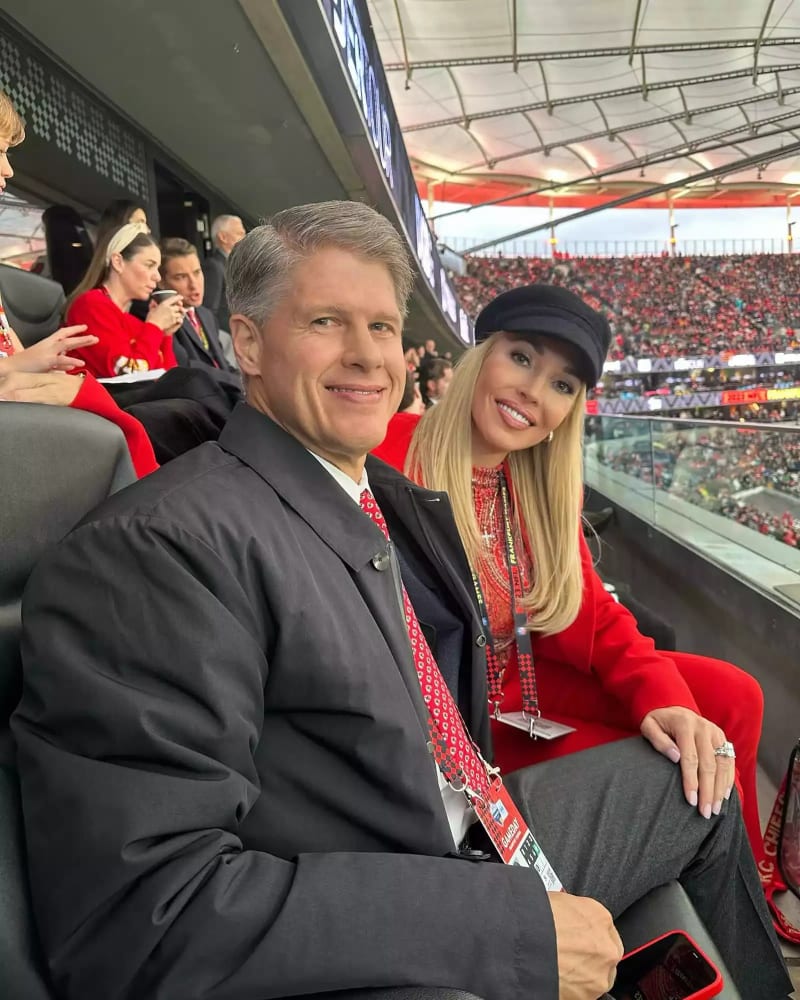 A man in a dark blazer and red patterned tie sits in a stadium suite box next to a woman with long blonde hair wearing a black baker boy cap and a red outfit, with a packed stadium visible in the background.