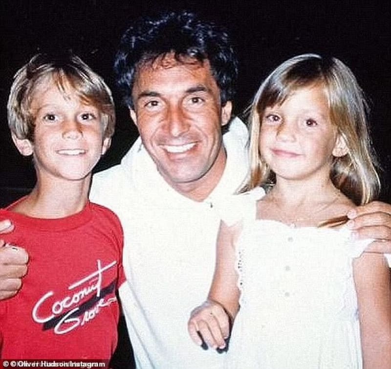 A man with dark hair wearing a white shirt stands smiling between two young blonde children — a boy in a red graphic T-shirt on the left and a girl in a white dress on the right — in an outdoor nighttime setting.