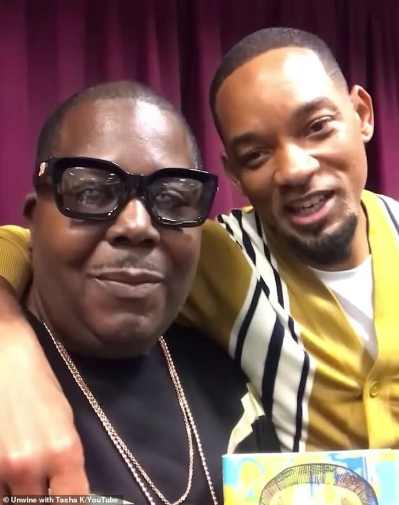 Two men posing closely together for a selfie-style photo in front of a dark red curtain backdrop, the man on the left wearing large black-framed glasses and a gold chain necklace, the man on the right wearing a yellow cardigan over a striped shirt with his arm around the other man.