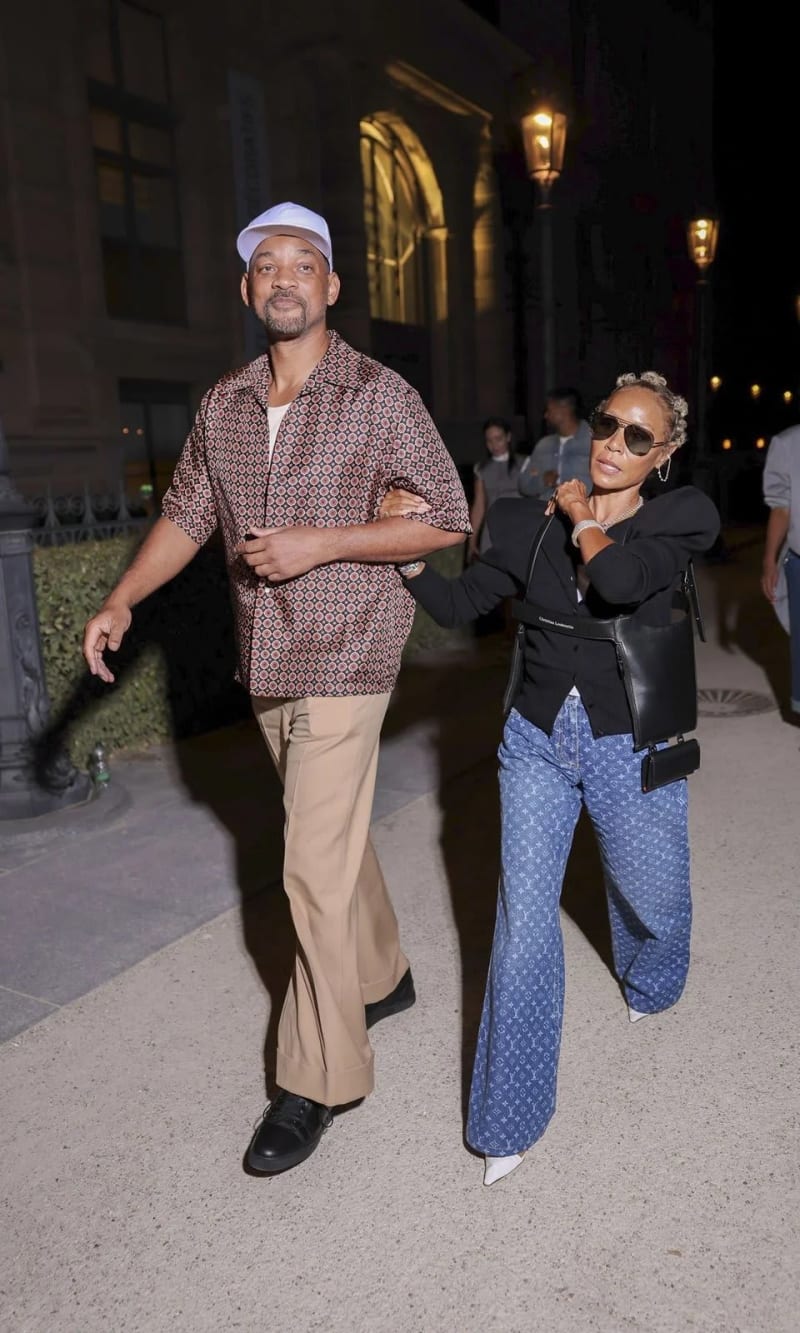 A tall man wearing a white baseball cap, a patterned short-sleeve button-up shirt, khaki wide-leg trousers, and black sneakers walks arm-in-arm with a woman wearing sunglasses, a black blazer, blue Louis Vuitton monogram wide-leg pants, and white heels, carrying a black leather crossbody bag, on a lit outdoor walkway at night.