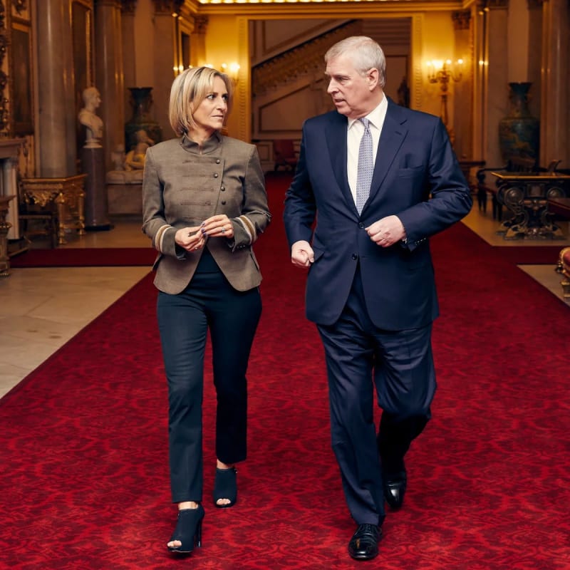 A woman with a blonde bob wearing a grey military-style jacket and black trousers walks alongside an older man in a navy suit and light blue tie across a red carpet in an ornate, gilded room.