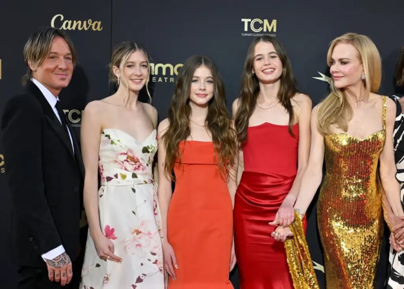 A man in a black suit stands with four women and girls posing together in front of a dark backdrop with Canva and TCM logos, wearing formal dresses in white floral, orange, red, and gold sequin.