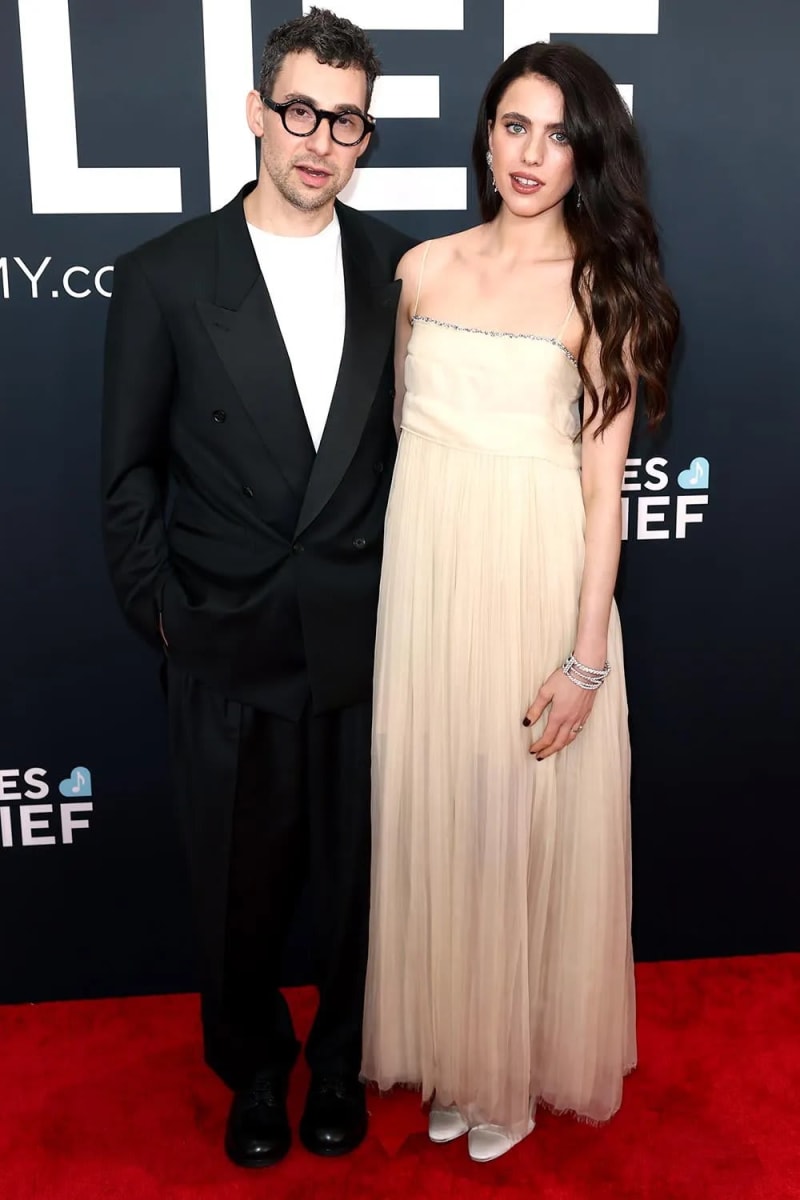 A man wearing black-framed glasses and a black double-breasted suit with a white t-shirt stands beside a woman with long dark wavy hair wearing a cream sleeveless tulle gown with crystal-trimmed straps, both standing on a red carpet in front of a dark branded backdrop.