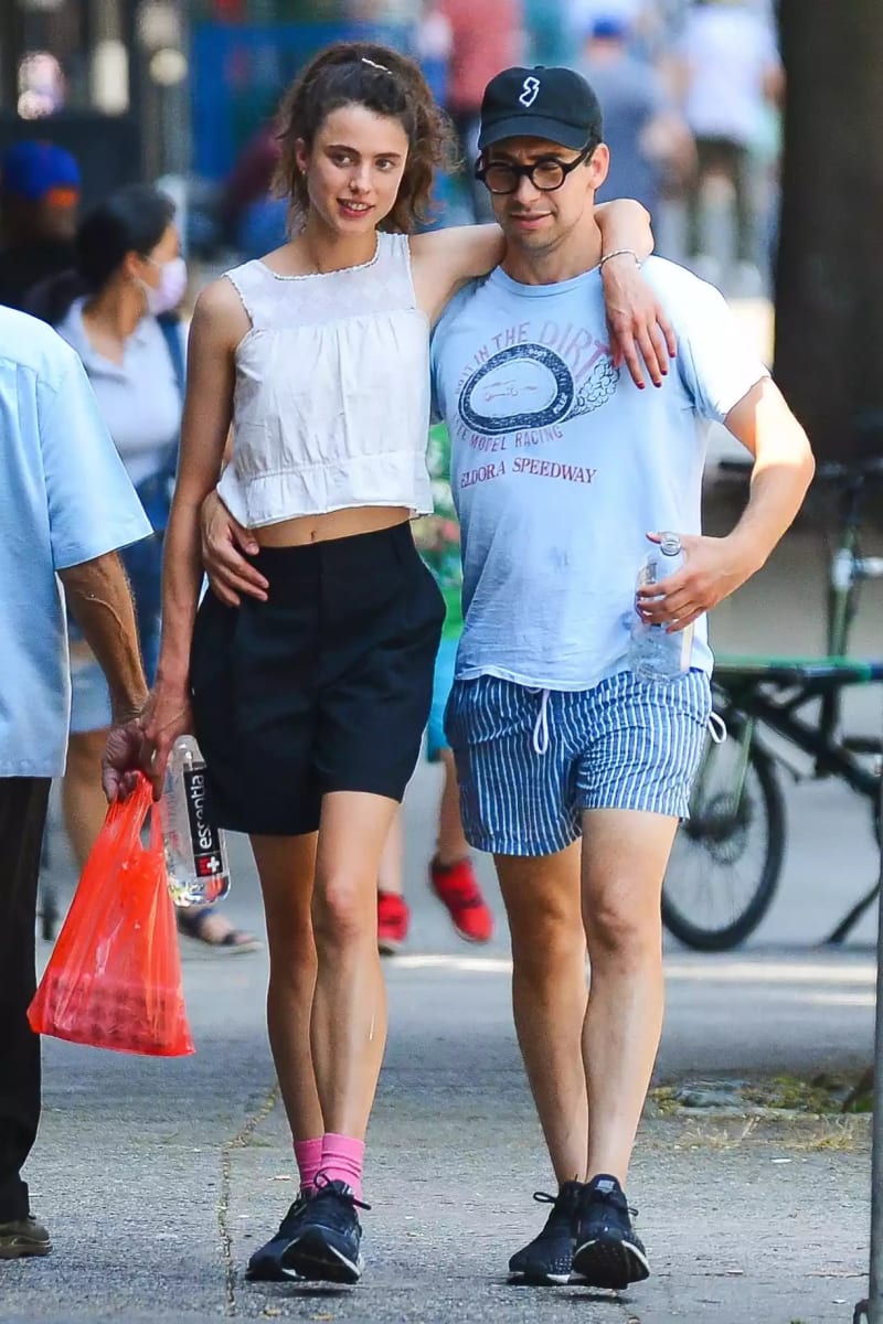 A tall woman in a white crop top, black biker shorts, pink socks, and black sneakers walks arm-in-arm with a man wearing a graphic t-shirt, blue striped shorts, a black cap, and glasses on a busy city sidewalk.
