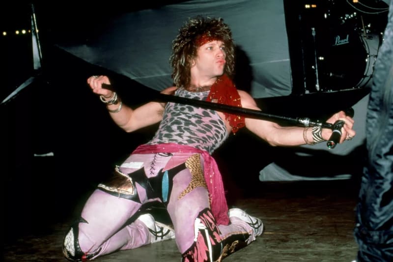 A man with a voluminous mullet and red bandana kneels on a dark stage, wearing a leopard-print sleeveless top, colorful patchwork pants, and sneakers, stretching a microphone stand horizontally across his body with both hands, with a Pearl drum kit visible in the background.