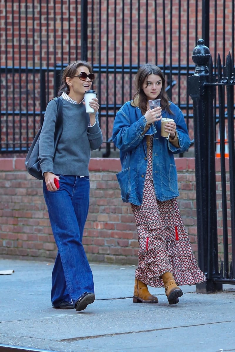 Two women walking side by side on a city sidewalk in front of a black iron fence and brick wall, both holding drinks — the woman on the left wearing sunglasses, a gray sweater, and wide-leg jeans carrying a white to-go cup, and the woman on the right wearing an oversized denim jacket over a long patterned dress with tan boots holding an iced drink.
