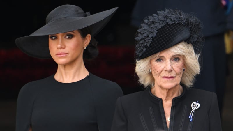 Two women standing side by side, both dressed in black formal attire and black hats, against a dark blurred backdrop.