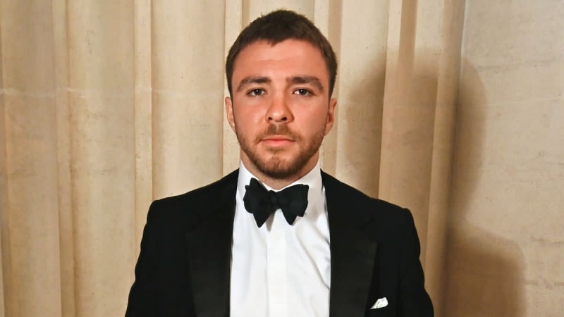 A man in a black tuxedo with a bow tie poses for a formal portrait against a beige curtained background.