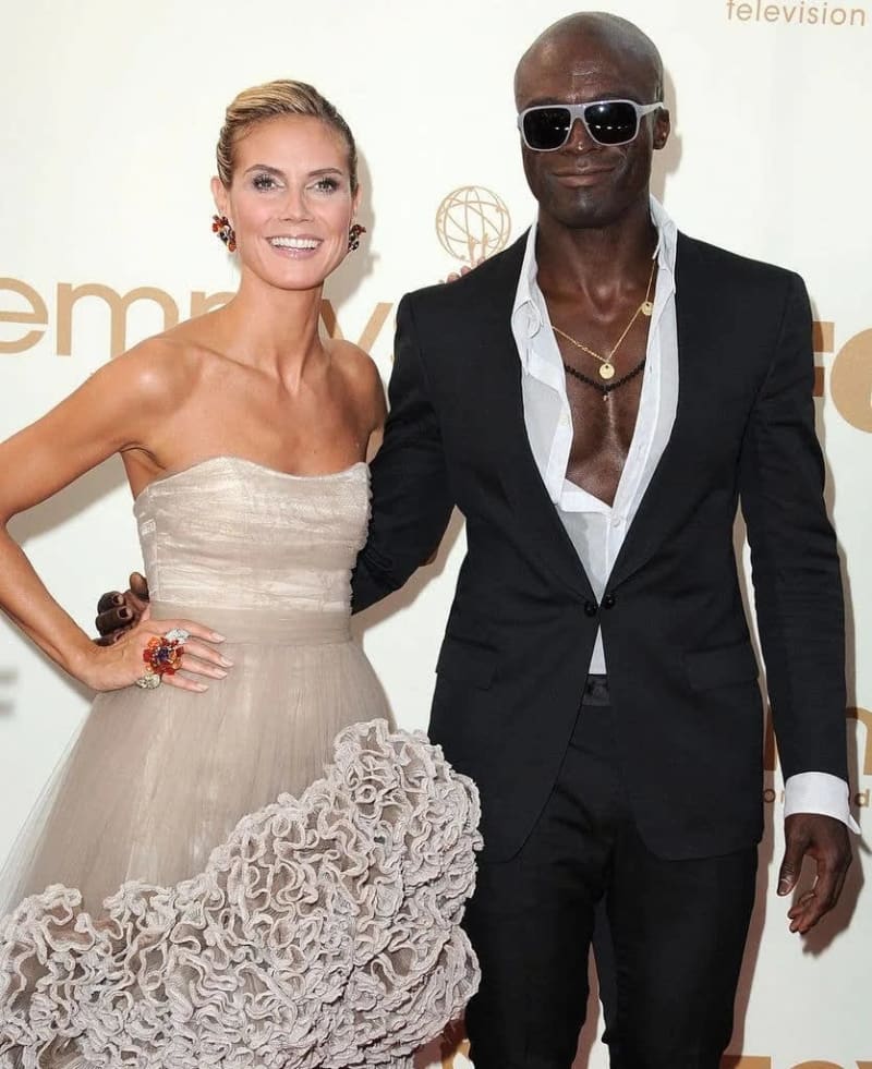 Two people at a formal event: a woman in a beige strapless gown and a man in a black suit with sunglasses.