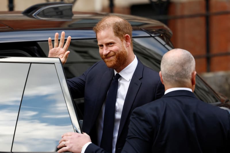 A man in a dark suit and tie waves while standing beside a car, greeting another man in formal attire near a brick building.